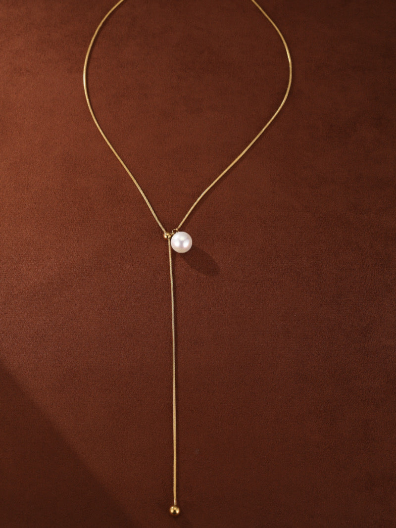 Sheen - Freshwater Pearl Long Necklace with a single 8mm round white freshwater pearl suspended on a delicate gold chain with an adjustable slider and a small gold bead at the end, on a brown background.