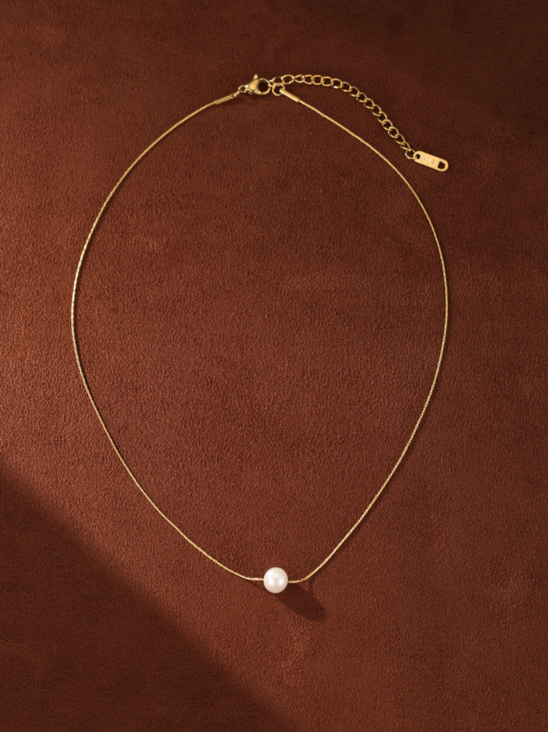 A delicate gold-toned chain necklace featuring a single, small, round white freshwater pearl pendant, displayed against a textured brown background.