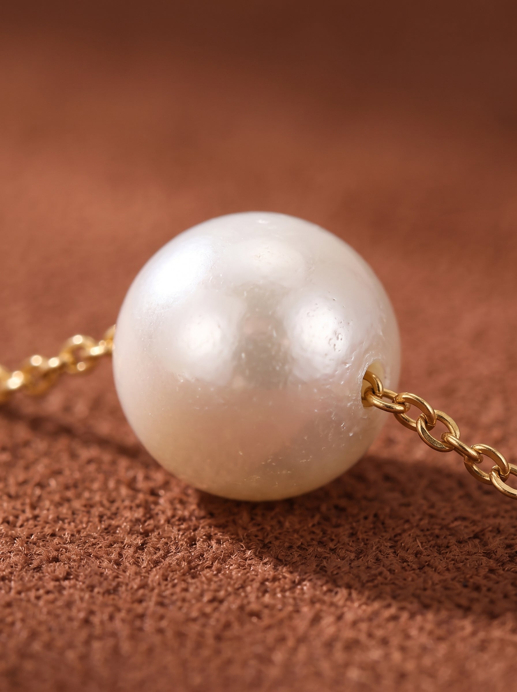 Close-up of the Solace pendant necklace featuring a single, lustrous, small white seed freshwater pearl on a delicate gold-toned chain against a textured brown background. @pearl