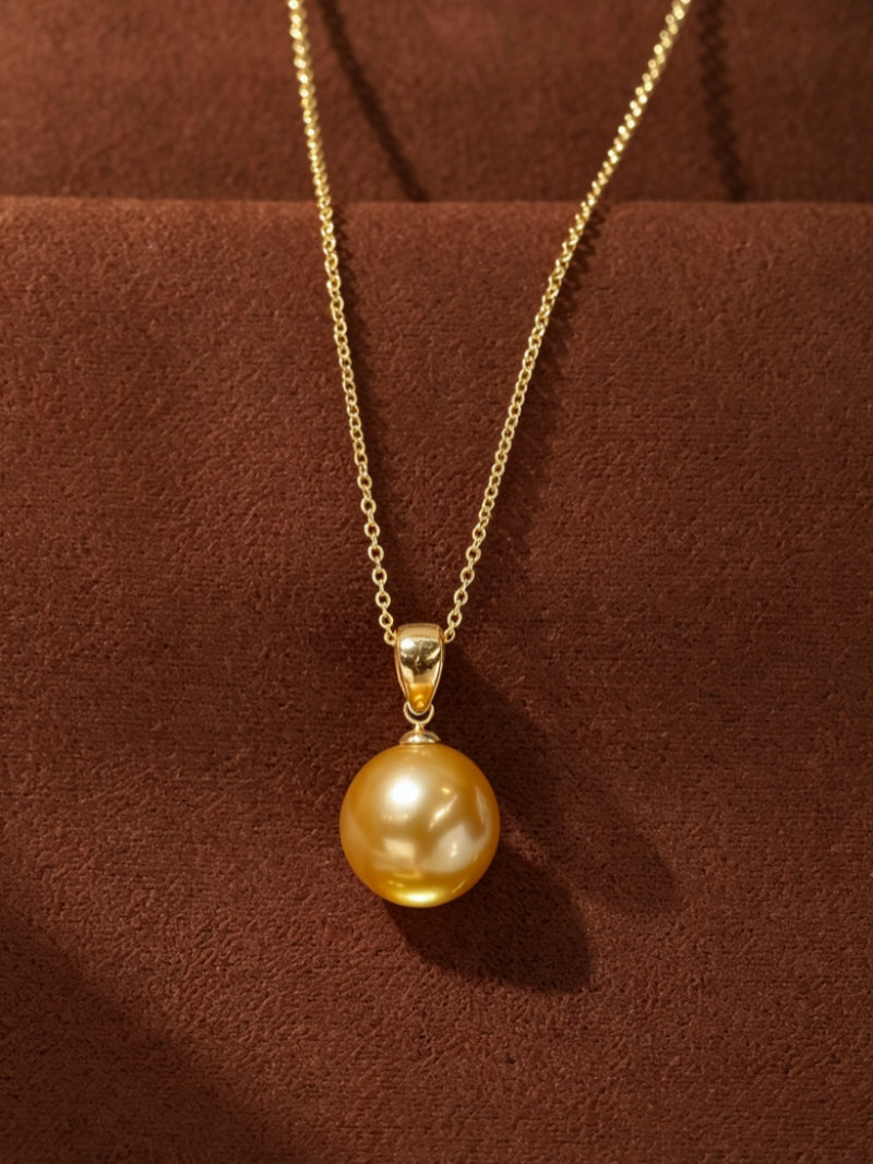 A close-up studio shot of the Solar Kiss pendant necklace, featuring a single, large, lustrous golden freshwater pearl suspended from a delicate gold-tone chain against a textured brown background.