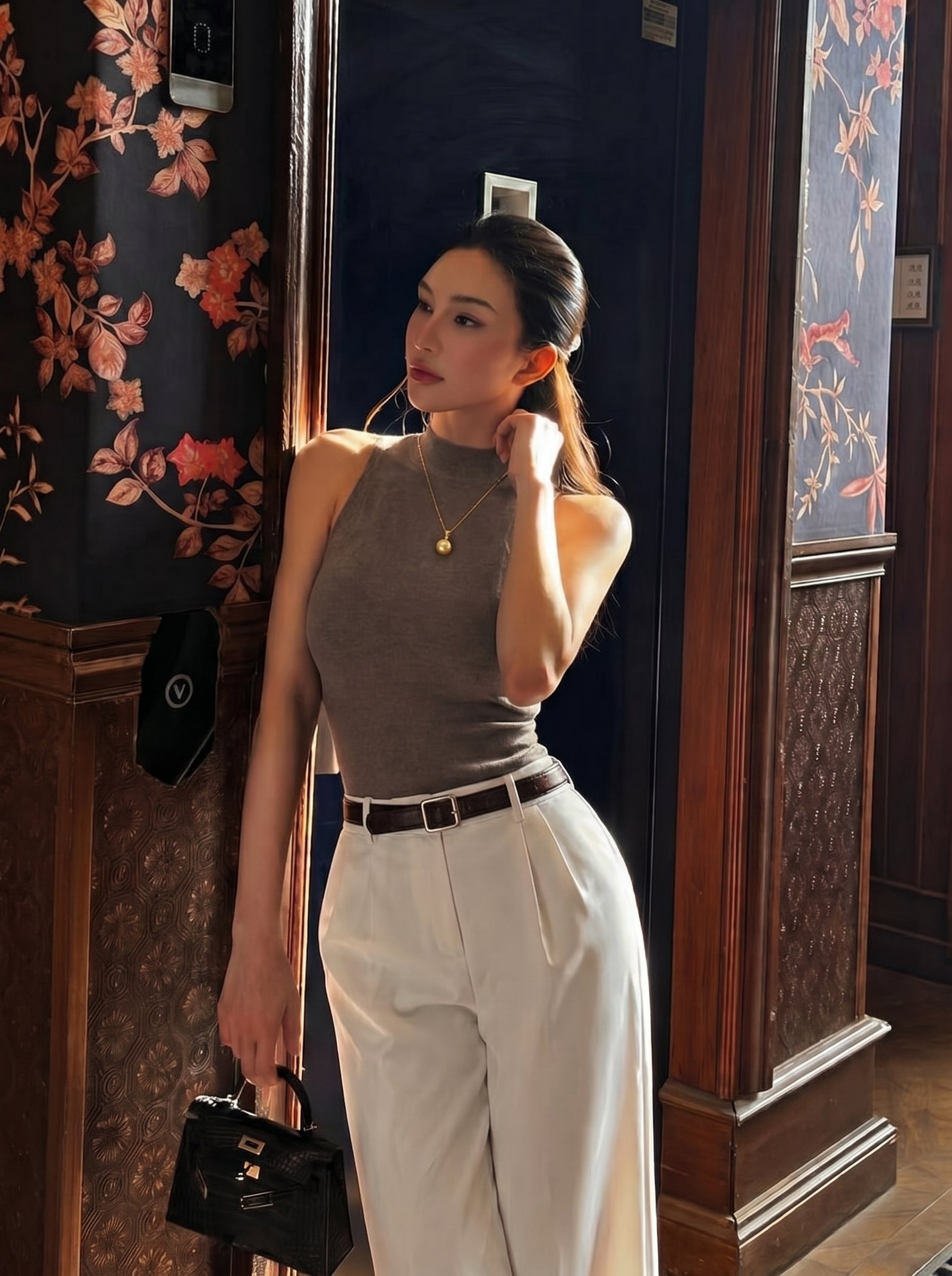 A woman wearing a grey sleeveless top, white pants, and a gold pendant necklace with a single round freshwater pearl, posing in a room with floral wallpaper and wooden accents. @lifestyle