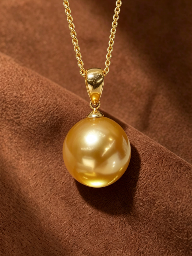 A close-up studio shot of the Solar Kiss necklace, featuring a single, lustrous 12mm round golden freshwater pearl suspended from a delicate gold-tone chain against a textured brown background.