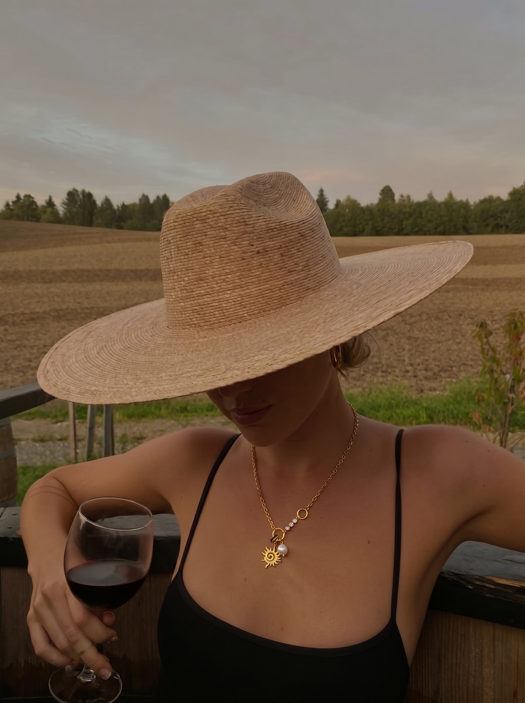 Woman wearing a Solaris freshwater pearl sun pendant necklace with a gold-tone chain and a white seed pearl, holding a glass of red wine in a vineyard setting. @lifestyle #lifestyle