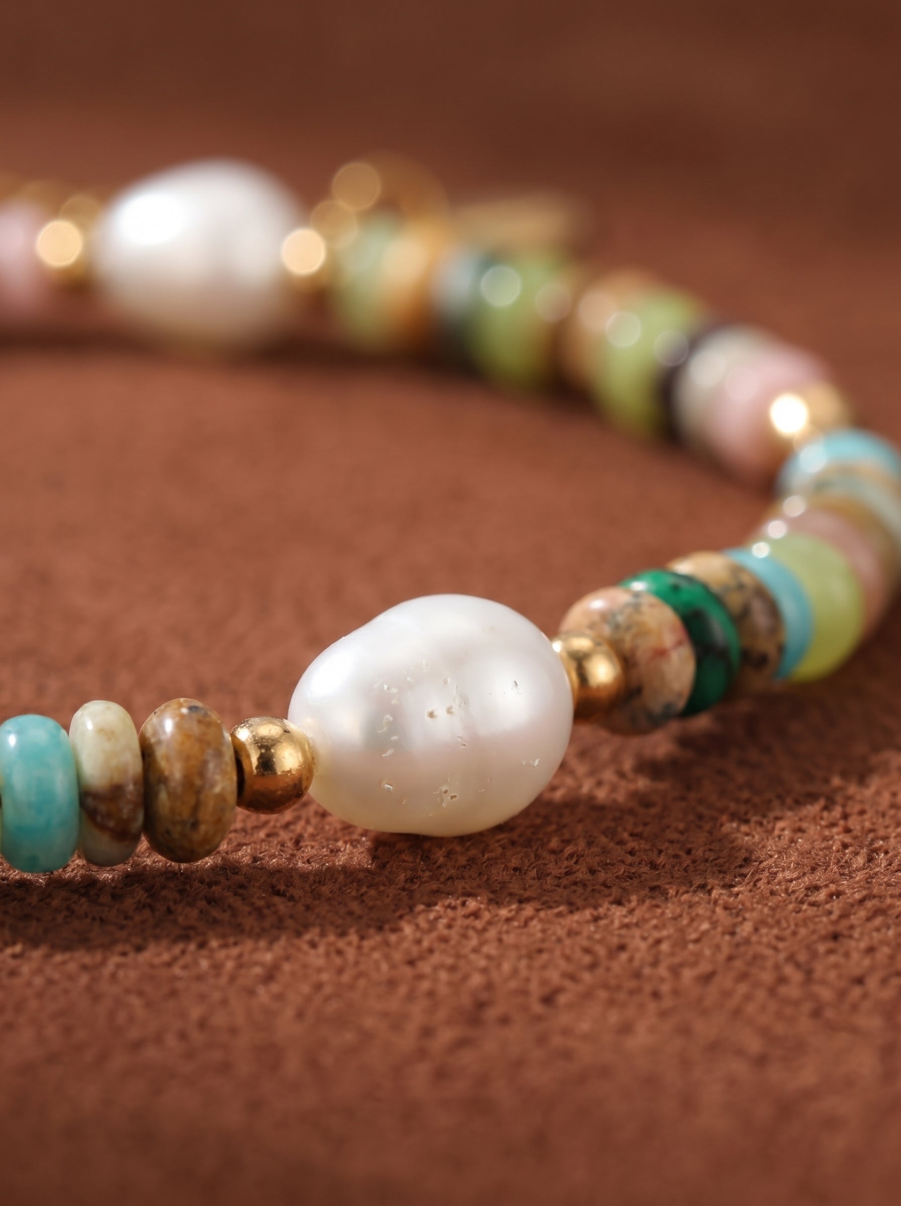 A close-up of the Solstice bracelet showcases a large, lustrous white freshwater pearl nestled amongst a colorful array of stone beads and gold spacers on a textured brown background. @pearl