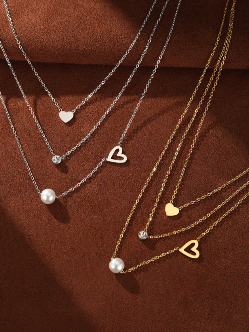 Un ensemble de collier superposé avec des chaînes délicates, présentant une seule perle blanche d'eau douce, une pierre accent scintillante, et des breloques en forme de cœur en tons argenté et doré.