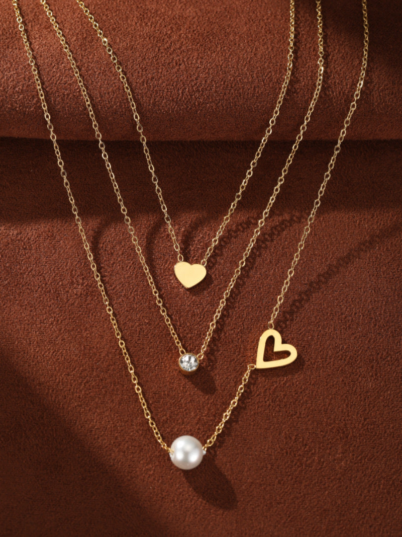 Collier superposé doré avec une perle d'eau douce, une pierre accent transparente, et deux breloques cœur sur fond texturé marron.