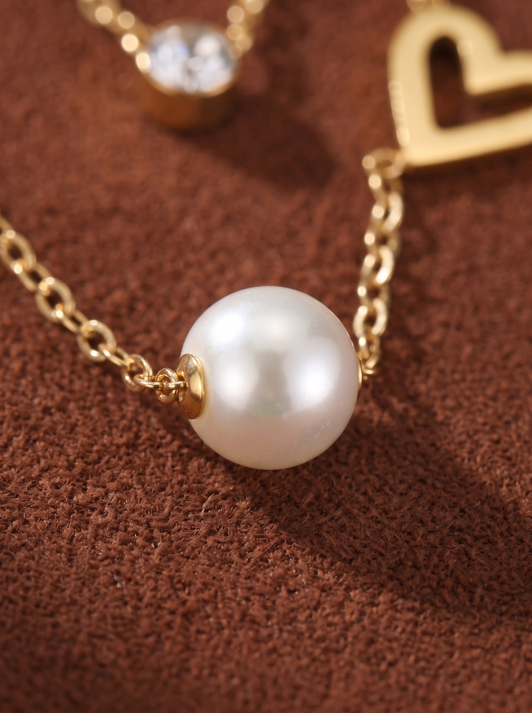 Gros plan d'un collier superposé doré avec une proéminente perle blanche d'eau douce de 4 mm, une discrète breloque cœur, et une pierre accent scintillante sur une chaîne délicate contre un fond texturé marron. @pearl