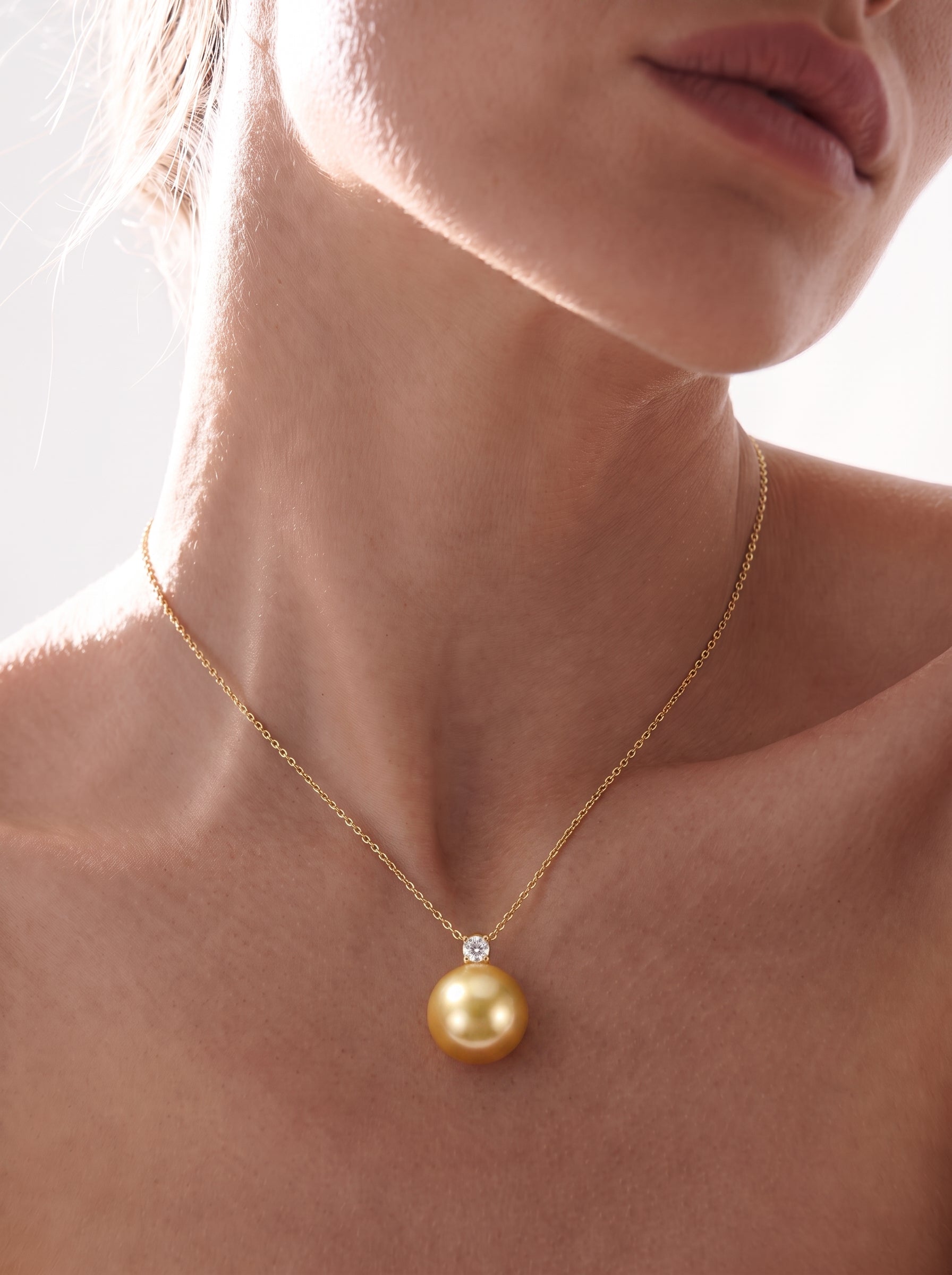 Close-up of a model wearing a Sovereign single pendant necklace featuring a luminous 9mm round golden South Sea pearl with a sparkling diamond accent on a delicate gold chain. @model