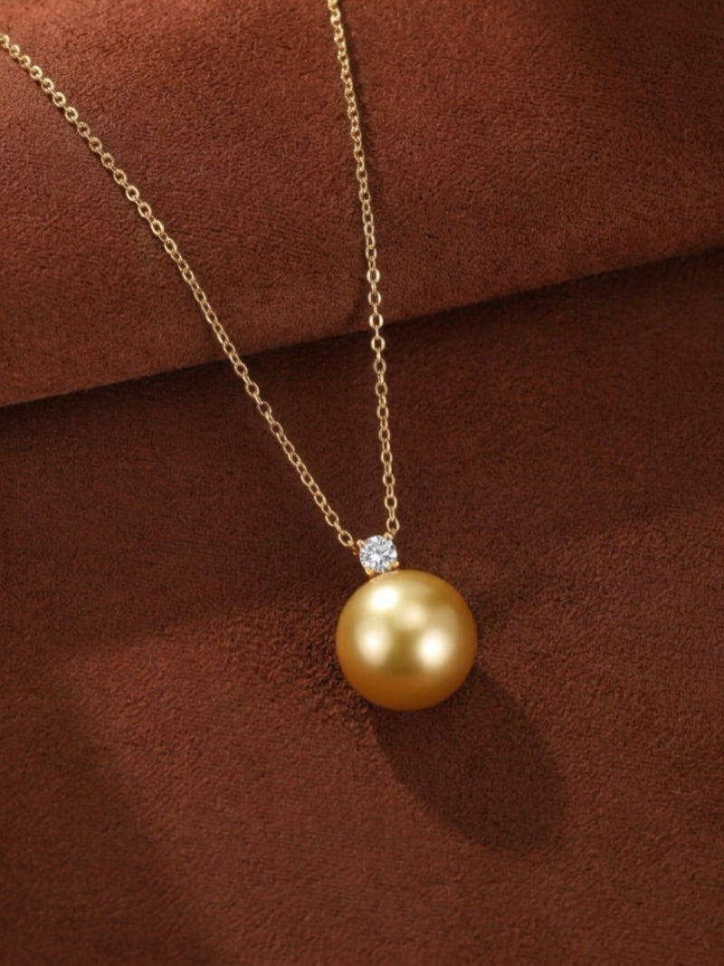 A single, luminous 9 mm round golden South Sea pearl is showcased on a delicate gold chain necklace, accented by a sparkling diamond. The pendant rests against a rich, textured brown background.