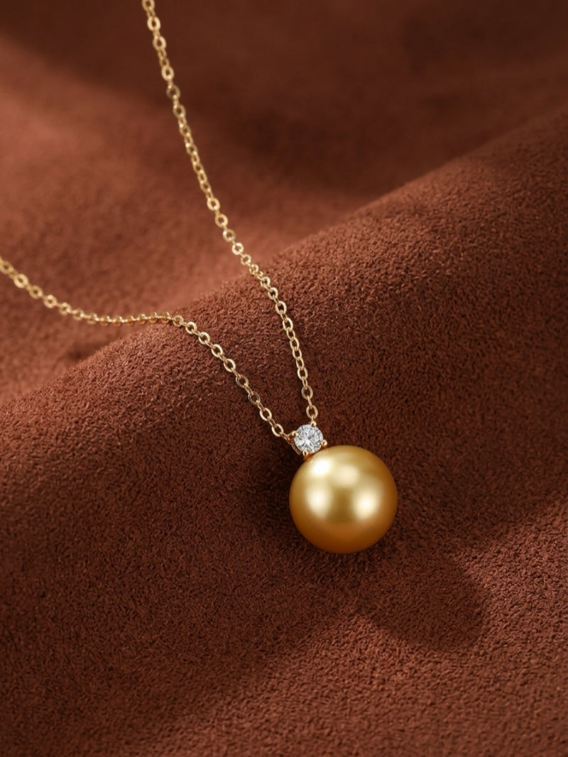 A single, lustrous 9mm round Golden South Sea Pearl pendant necklace, accented by a sparkling diamond, hangs from a delicate gold chain on a textured brown background.