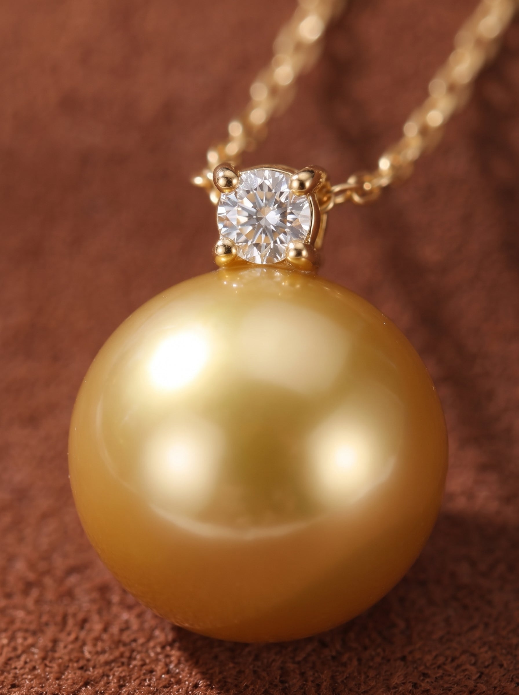A close-up of a pendant necklace featuring a luminous 9mm round golden South Sea pearl suspended below a sparkling diamond accent, on a delicate gold chain against a textured brown background. @pearl