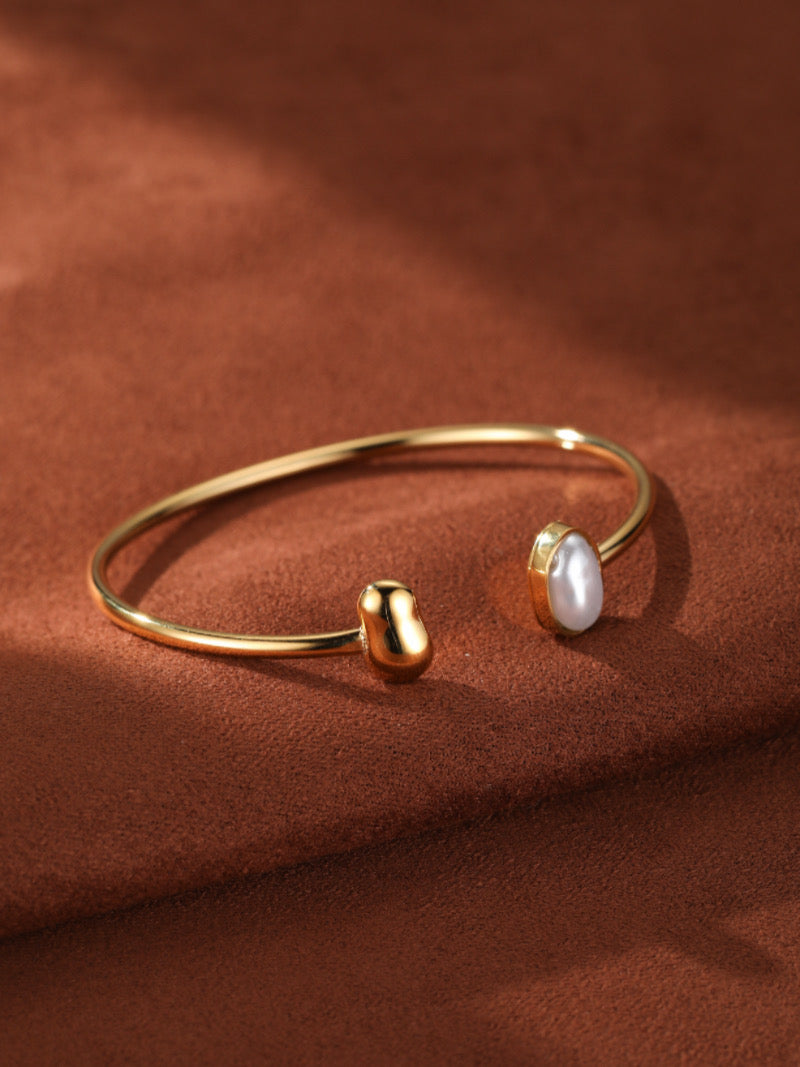 Gold cuff bracelet with a freshwater pearl on one end and a smooth gold sphere on the other, set against a textured brown background.
