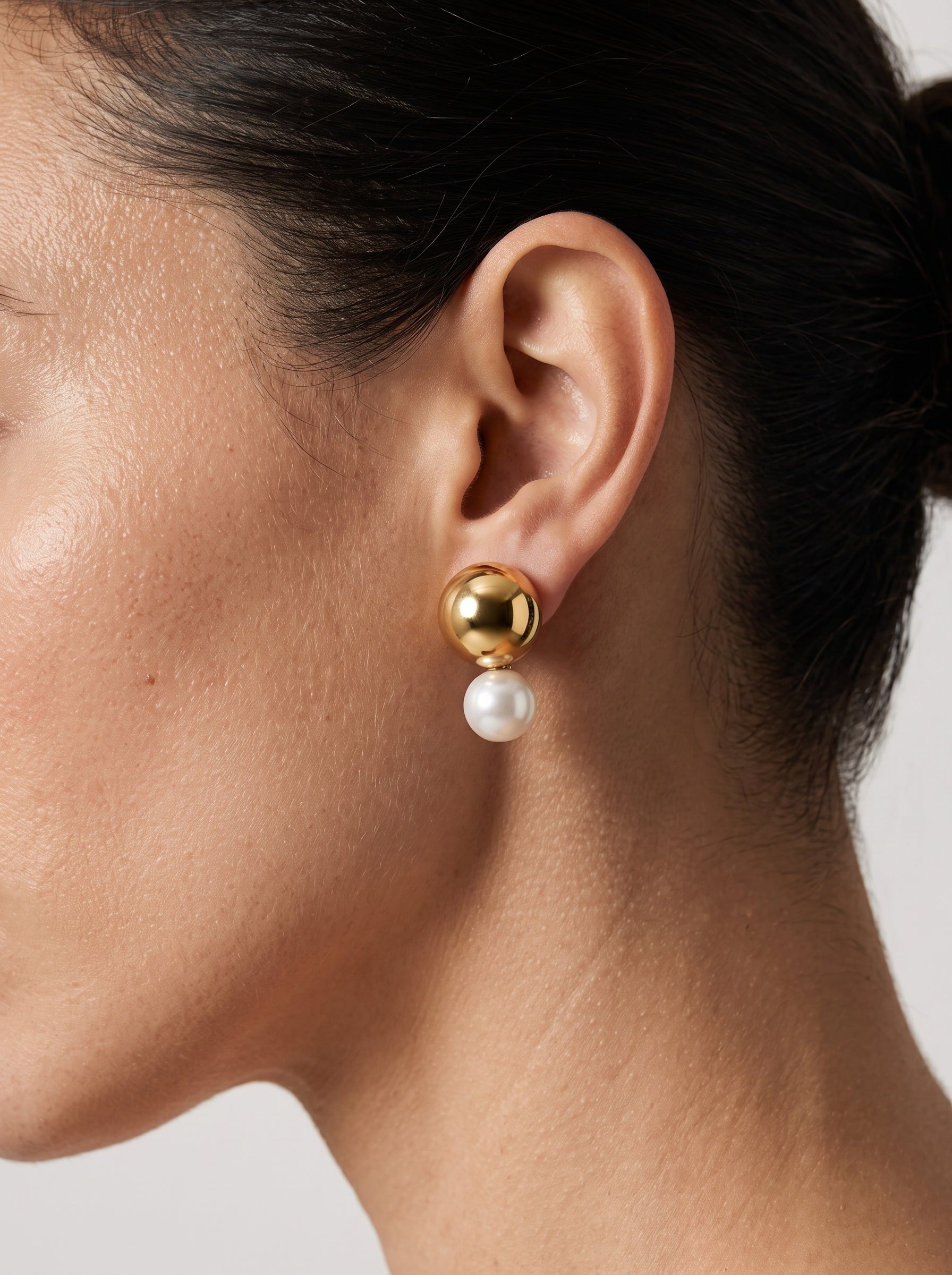 A close-up shot of a model wearing Sphere - Freshwater Pearl Stud Earrings, featuring a gold-toned sphere and a luminous white freshwater pearl. @model