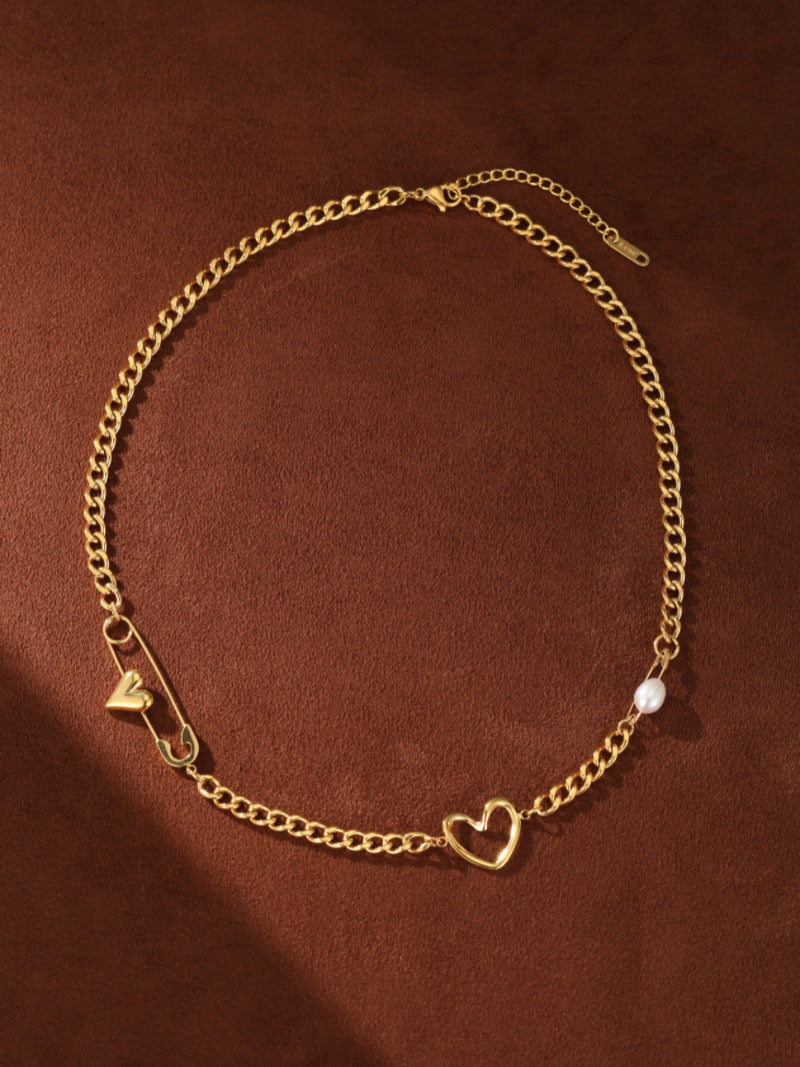 Collier ras-de-cou à chaîne gourmette imposante ton or avec une breloque épingle de sûreté, une petite breloque cœur, une breloque cœur ouvert, et une seule perle blanche d'eau douce.