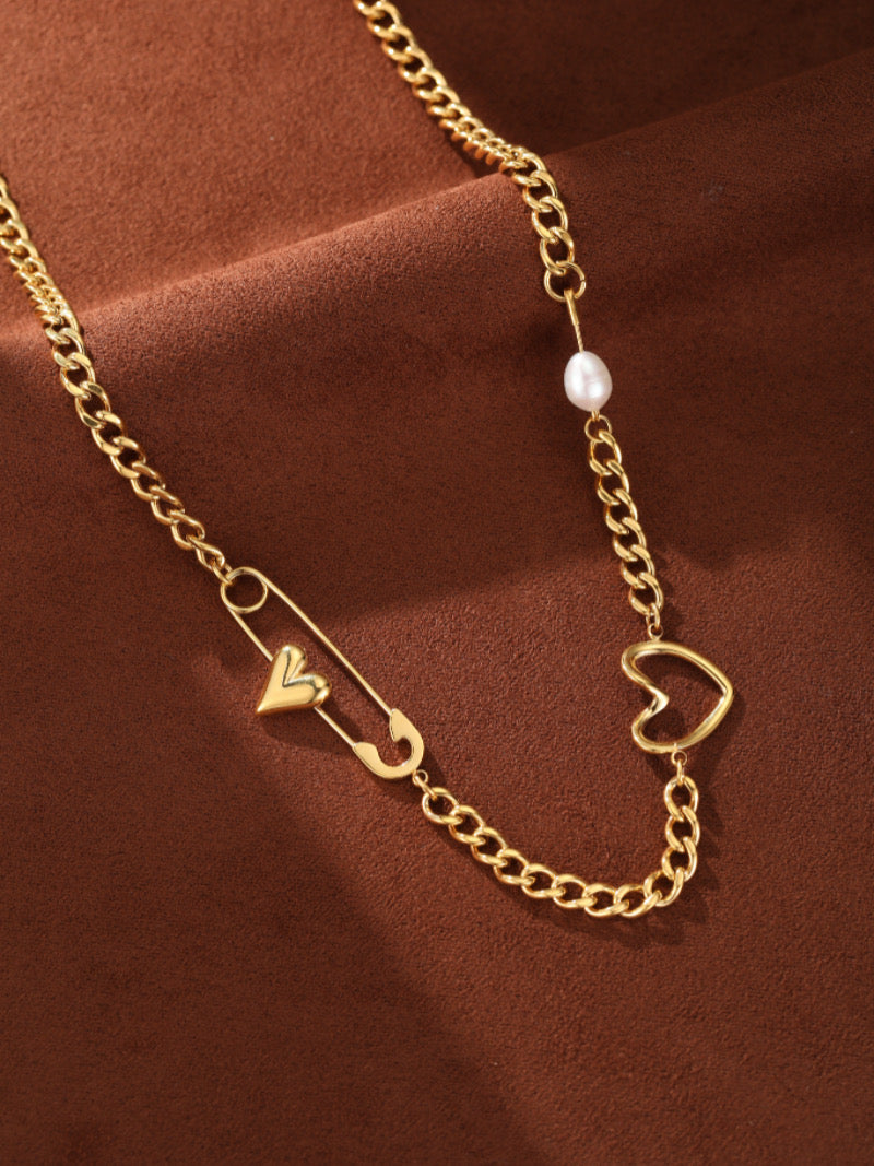 Collier à chaîne gourmette imposante ton or présentant une breloque épingle de sûreté avec un petit cœur, une plus grande breloque cœur ouvert, et une seule perle d'eau douce.