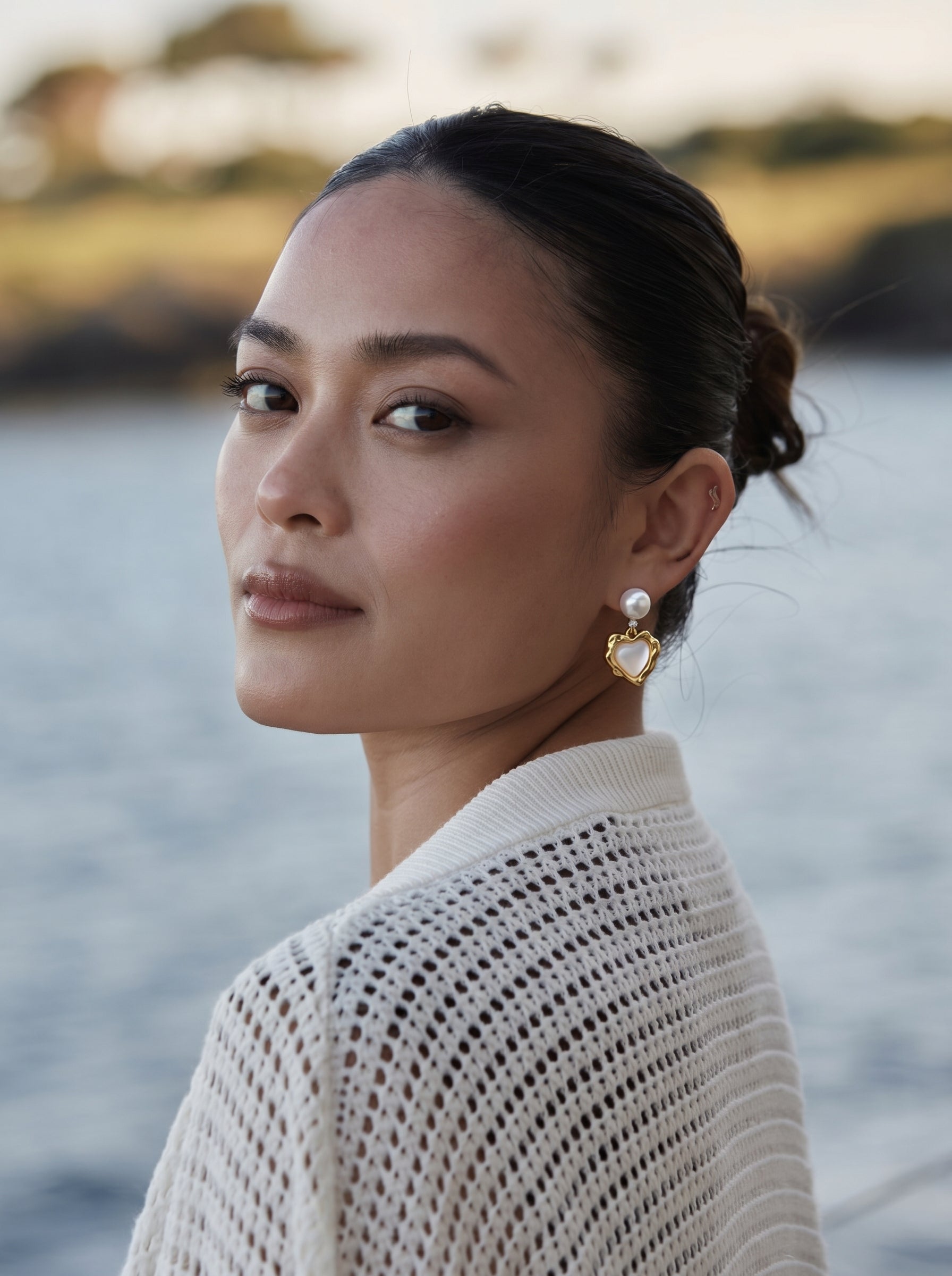 A woman wearing Starlight Freshwater Pearl Star Stud Drop Earrings, featuring a white pearl stud and a gold-tone heart motif, with a blurred water background. @lifestyle #lifestyle