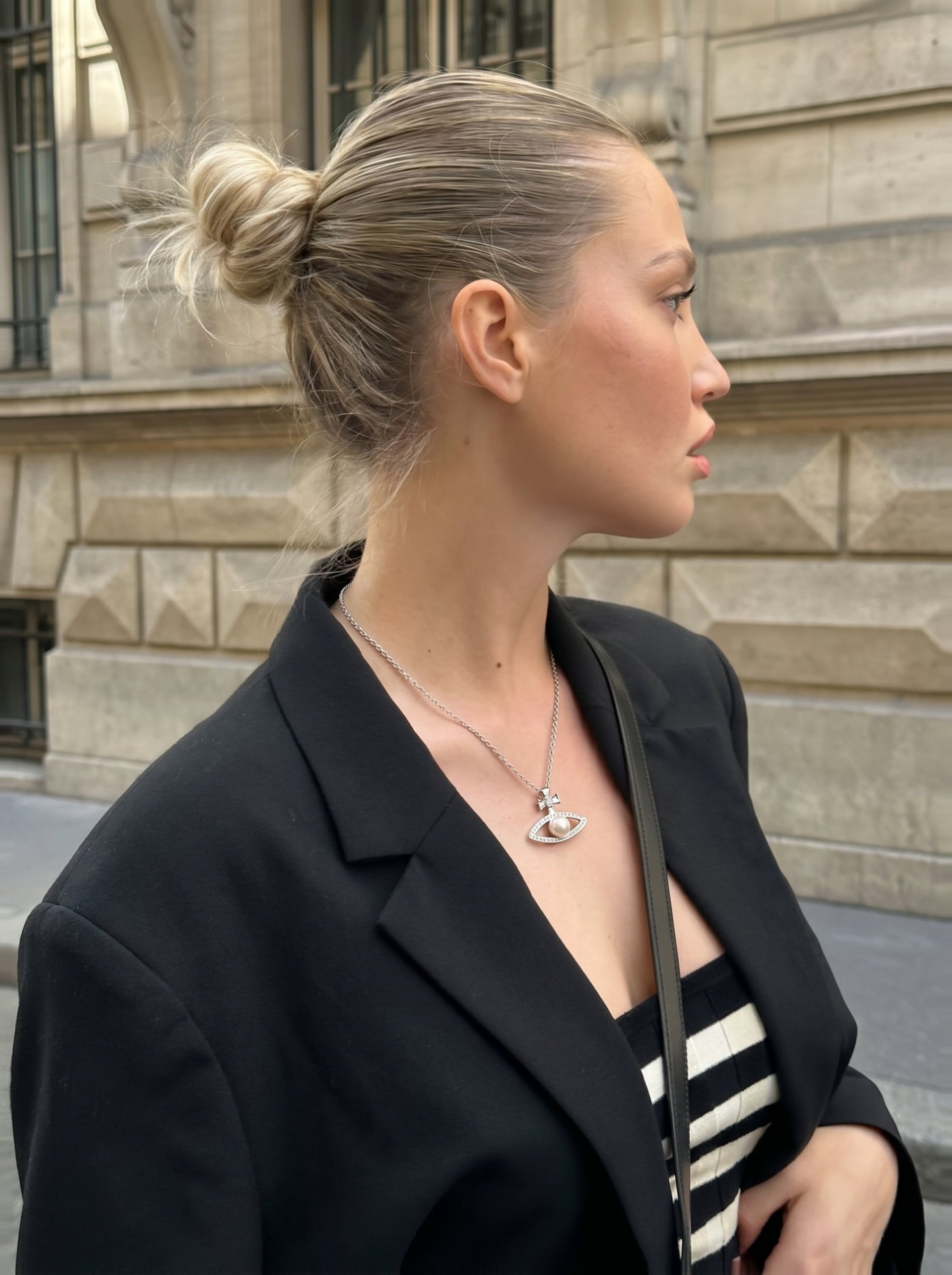A woman with her hair in a bun wears the Stellar Gaze Freshwater Pearl Cross Chain Pendant Necklace, featuring an eye-shaped pendant with a white pearl and sparkling accents, topped with a cross motif, layered over a striped top and black blazer. @lifestyle #lifestyle