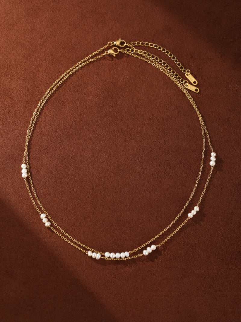 A delicate double-strand gold-toned necklace featuring small, luminous freshwater pearls spaced along the chains against a brown background.