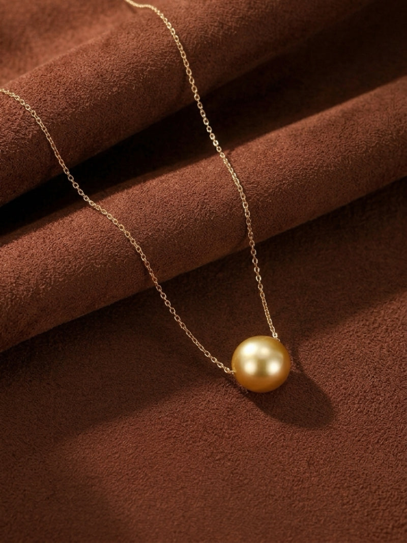 A single, lustrous 9-10 mm golden South Sea pearl is suspended from a delicate, fine gold chain. The necklace is displayed against a textured brown background, highlighting the pearl's natural luster and the minimalist design. Sunbeam - South Sea Pearl Single Pendant Necklace.