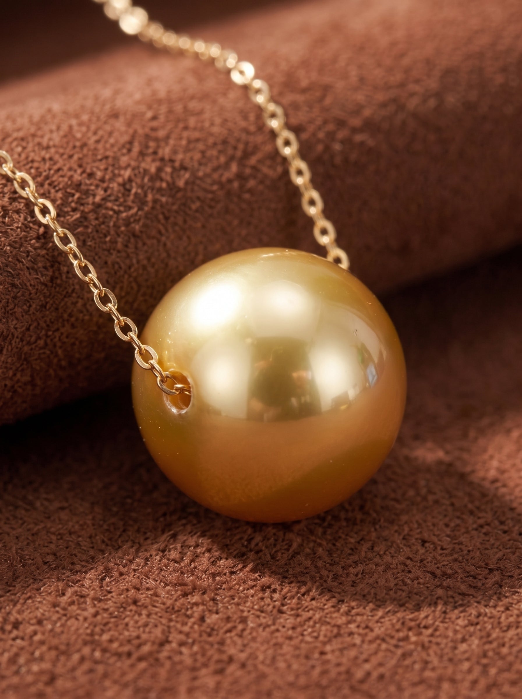 A close-up shot of a single, lustrous 9-10mm golden South Sea pearl pendant necklace with a delicate gold chain, resting on a textured brown surface. @pearl