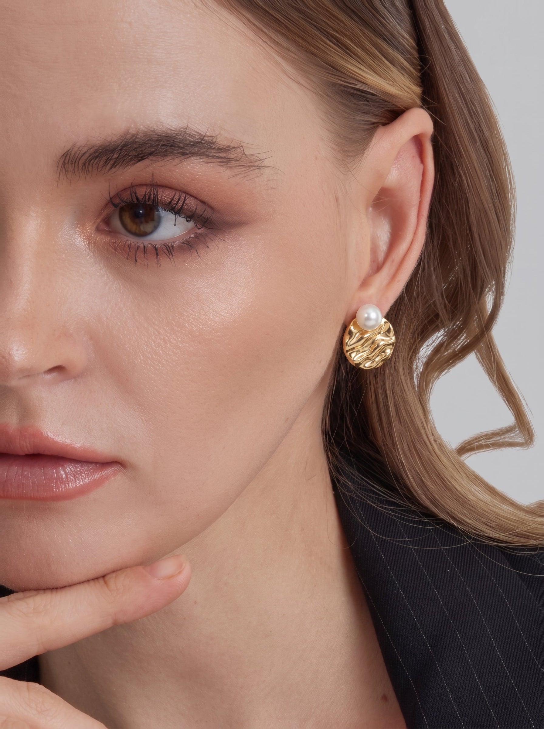 Gros plan d'une femme portant des boucles d'oreilles puces ton or avec un disque texturé façon vague et une petite perle blanche d'eau douce au centre. @model
