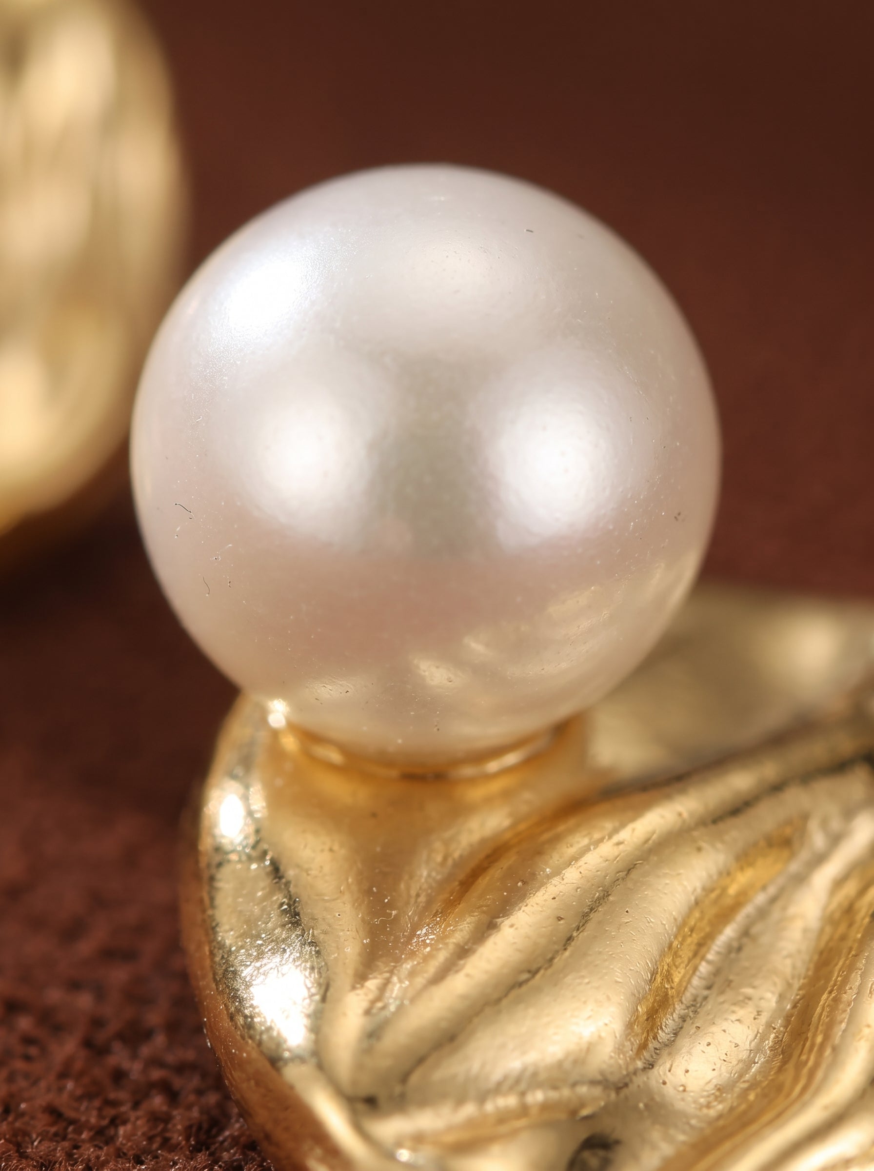 Gros plan d'une seule boucle d'oreille puce Surge en perle d'eau douce, présentant une lustrée perle ronde blanche de 4 mm au sommet d'un disque ton or au design texturé et ondulé. @pearl