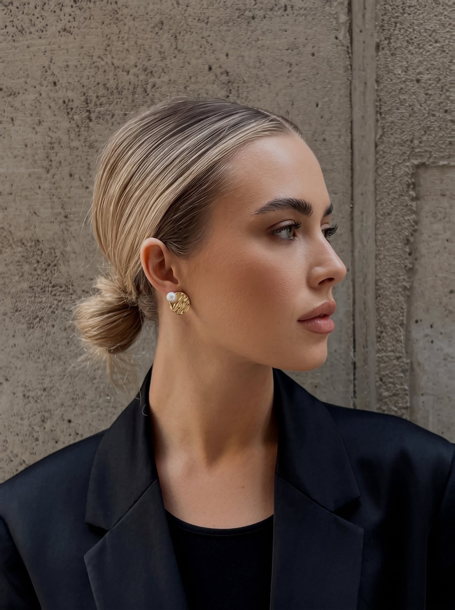 Une femme portant un blazer noir et des boucles d'oreilles puces ton or avec des perles blanches d'eau douce se tient contre un mur en béton texturé. @lifestyle
