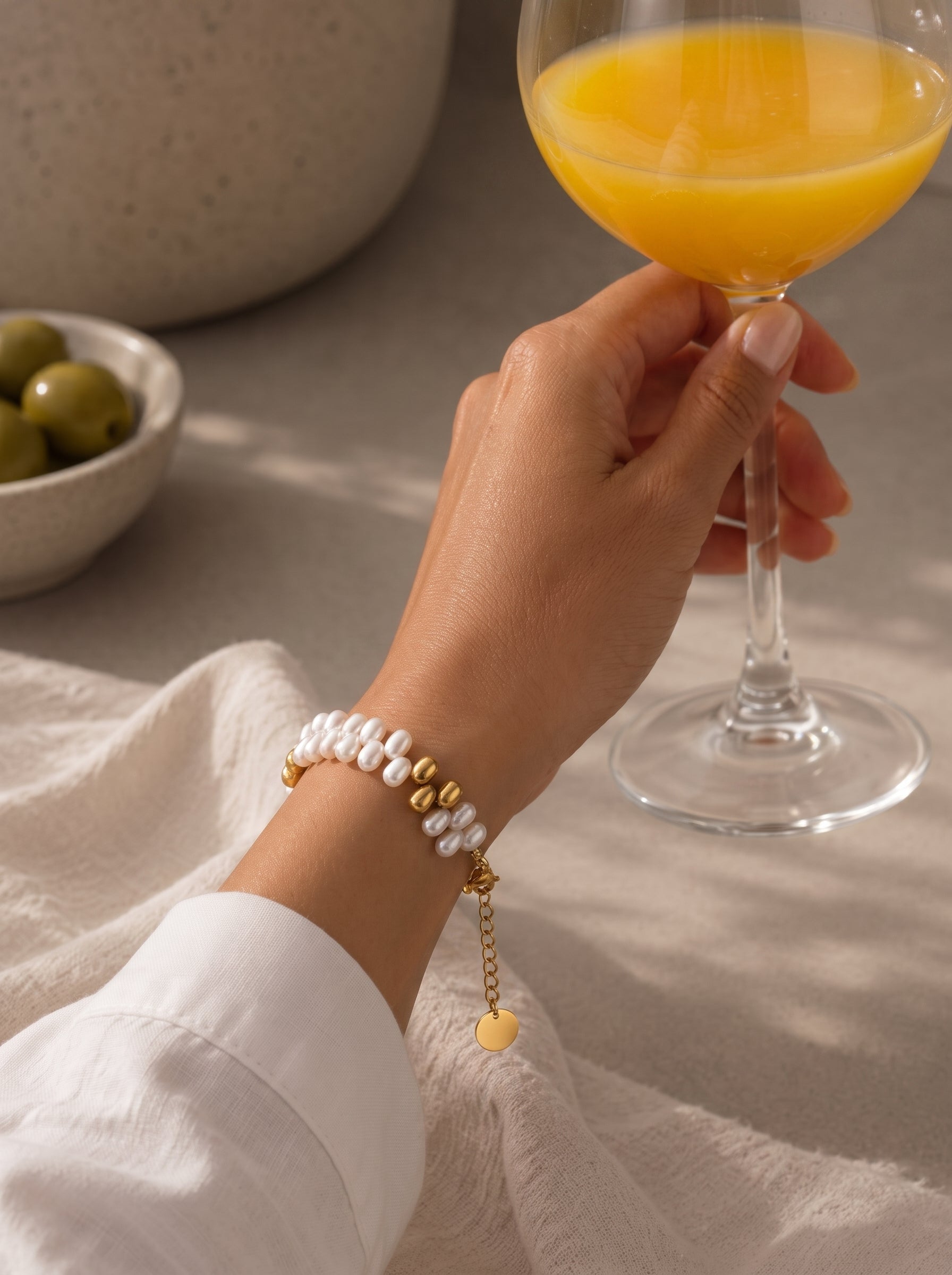 A woman's wrist wearing the Sylvan Freshwater Pearl Beaded Bracelet, featuring white pearls and gold-toned beads, while holding a glass of orange juice. @lifestyle