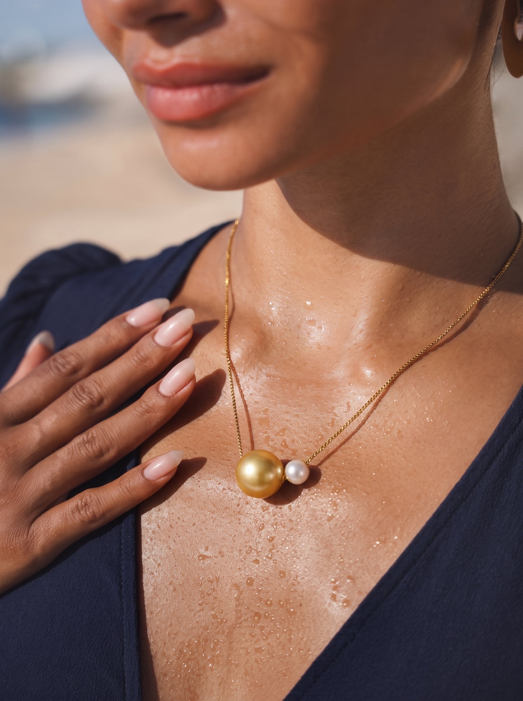 A close-up shot of a woman wearing a gold pendant necklace featuring a large golden South Sea pearl and a smaller white Akoya pearl, with a beachy background. @lifestyle