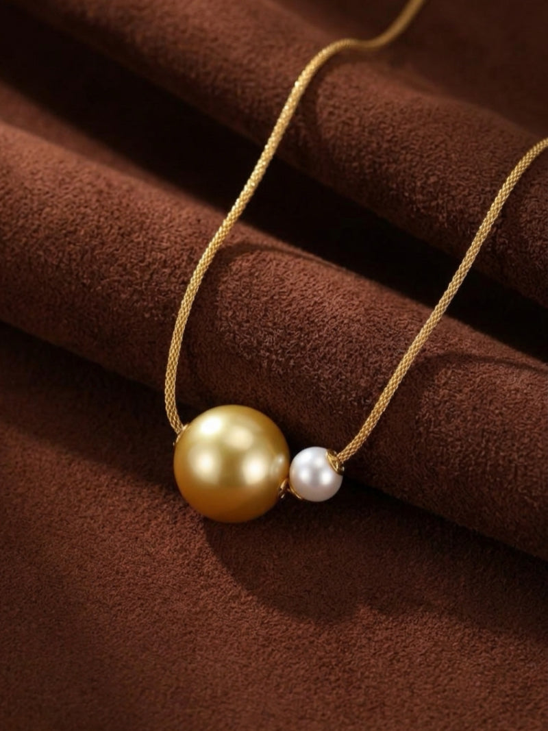 A delicate gold chain holds a large, lustrous golden South Sea pearl next to a smaller, radiant white Akoya pearl, displayed on a textured brown background.