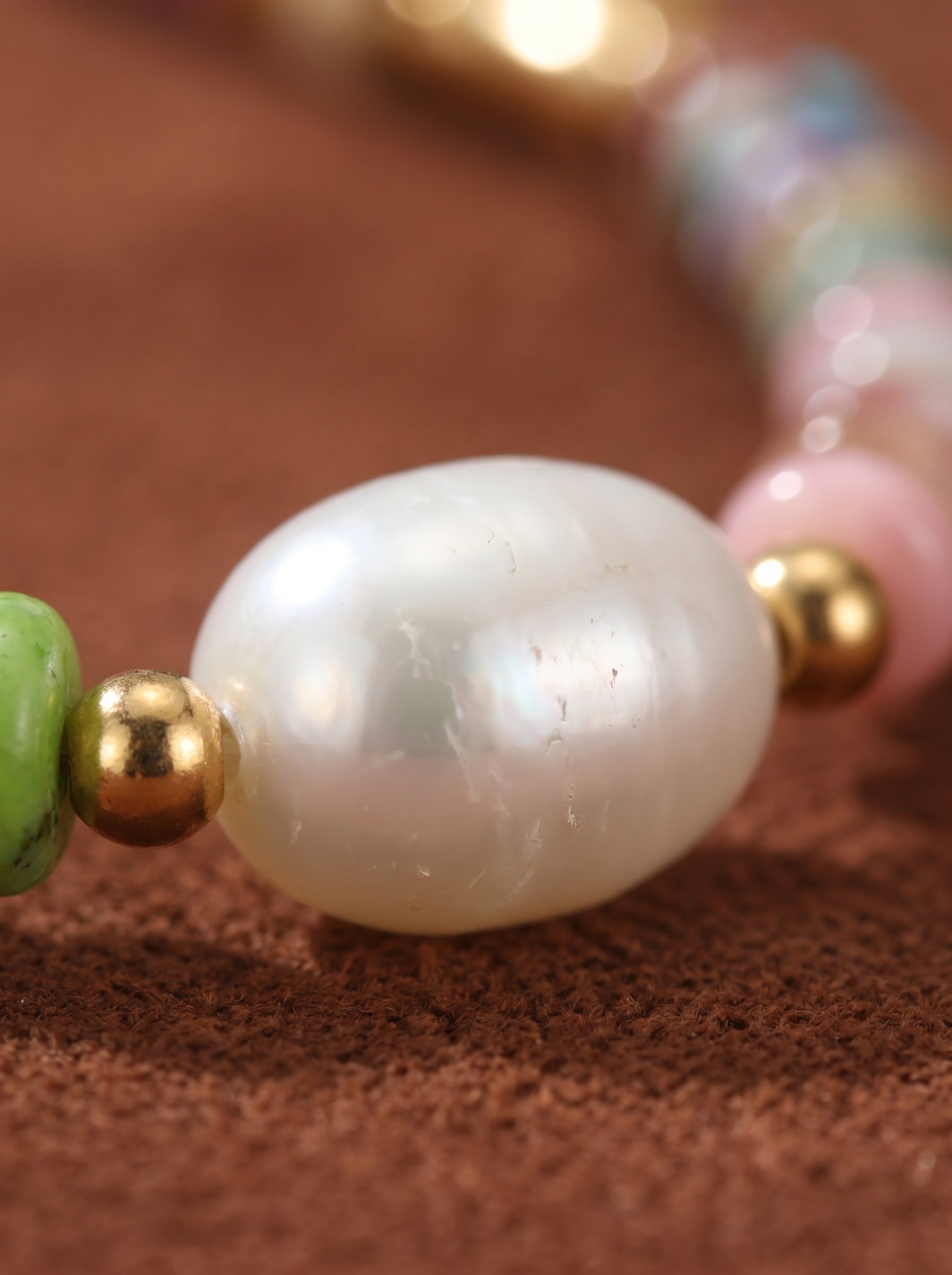 Close-up of a Tapestry necklace featuring a large, irregular freshwater pearl, interspersed with colorful beads and gold accents on a brown textured background. @pearl