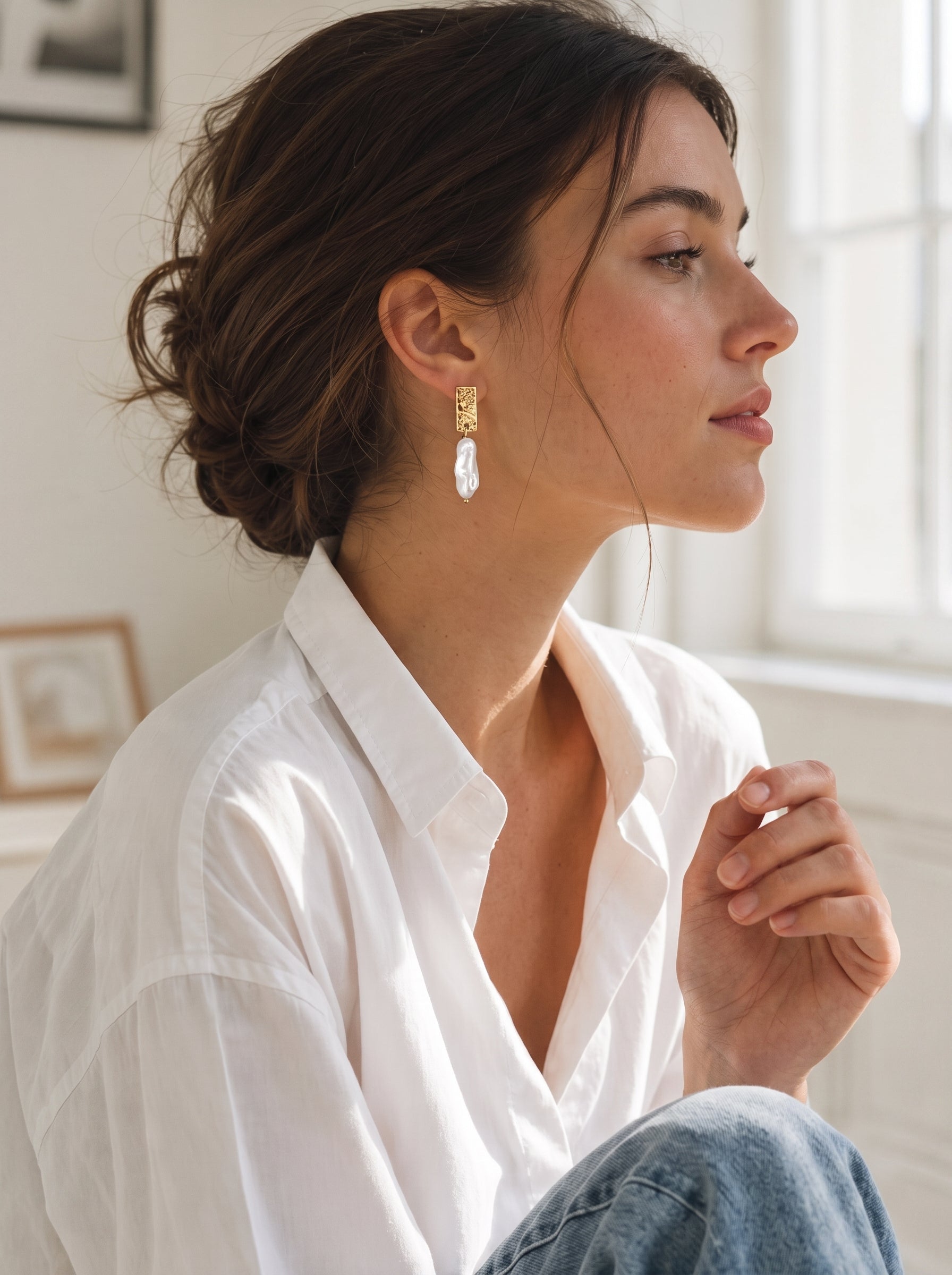 Femme portant les boucles d'oreilles goutte en perle d'eau douce Terra avec une puce martelée dorée et une goutte de perle baroque blanche organique, dans un cadre lumineux et naturel. @lifestyle
