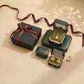 A collection of dark green vintage-style jewelry boxes, one open to reveal a pearl pendant necklace, another open to show pearl earrings, and a closed black gift box tied with a burgundy ribbon. @shared