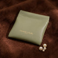 A soft, sage green magnetic pochette with gold embossed "Grande Lilith" logo sits on a brown textured surface next to a few pearls. @shared