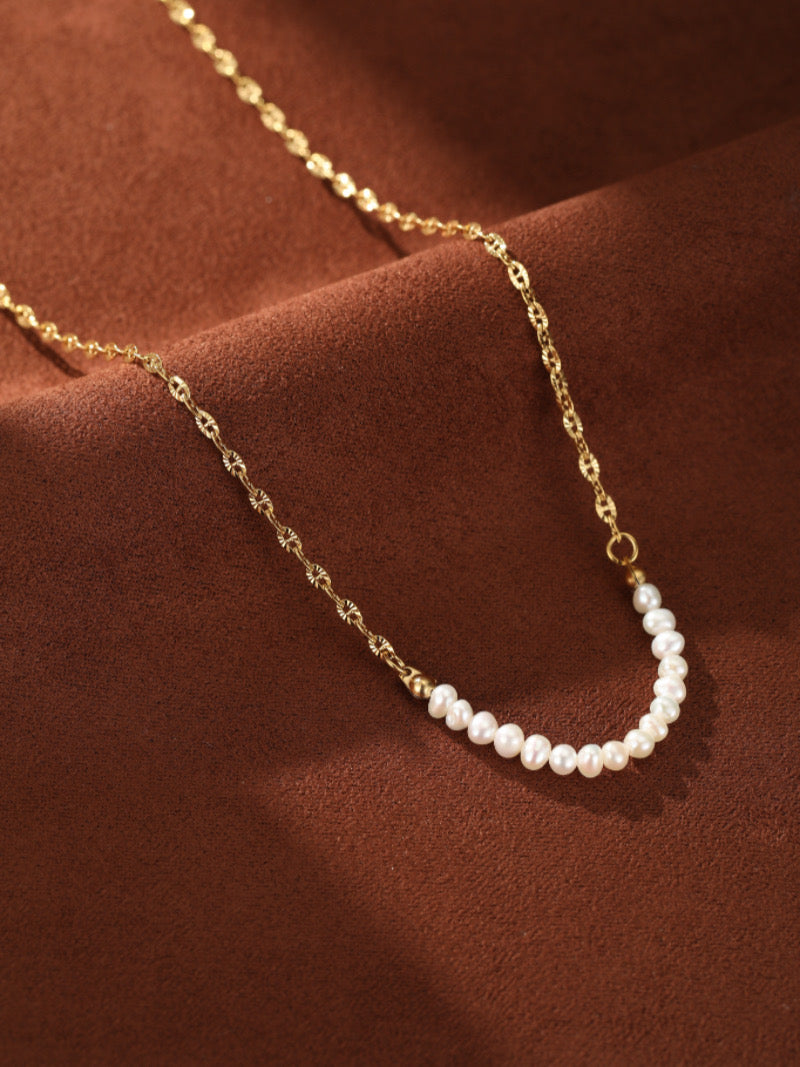 Trace - Collier à chaîne ton or présentant un délicat arc de petites et lustrées perles blanches d'eau douce sur fond texturé marron.