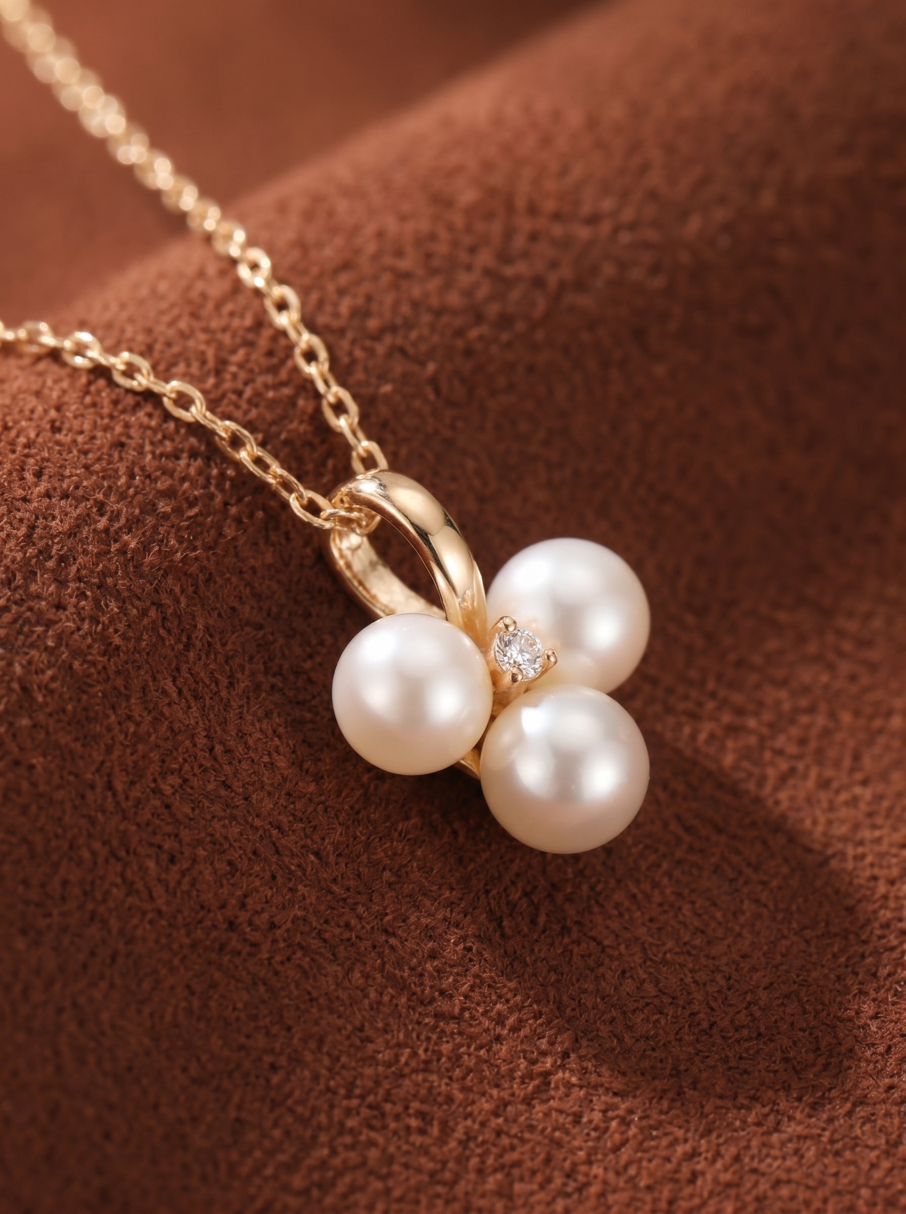 A close-up shot of the Trine Freshwater Pearl Trio Pendant Chain Necklace, showcasing three lustrous 6mm white pearls clustered together on a delicate gold-tone chain, accented by a single sparkling stone. The pendant is set against a textured brown background. @pearl