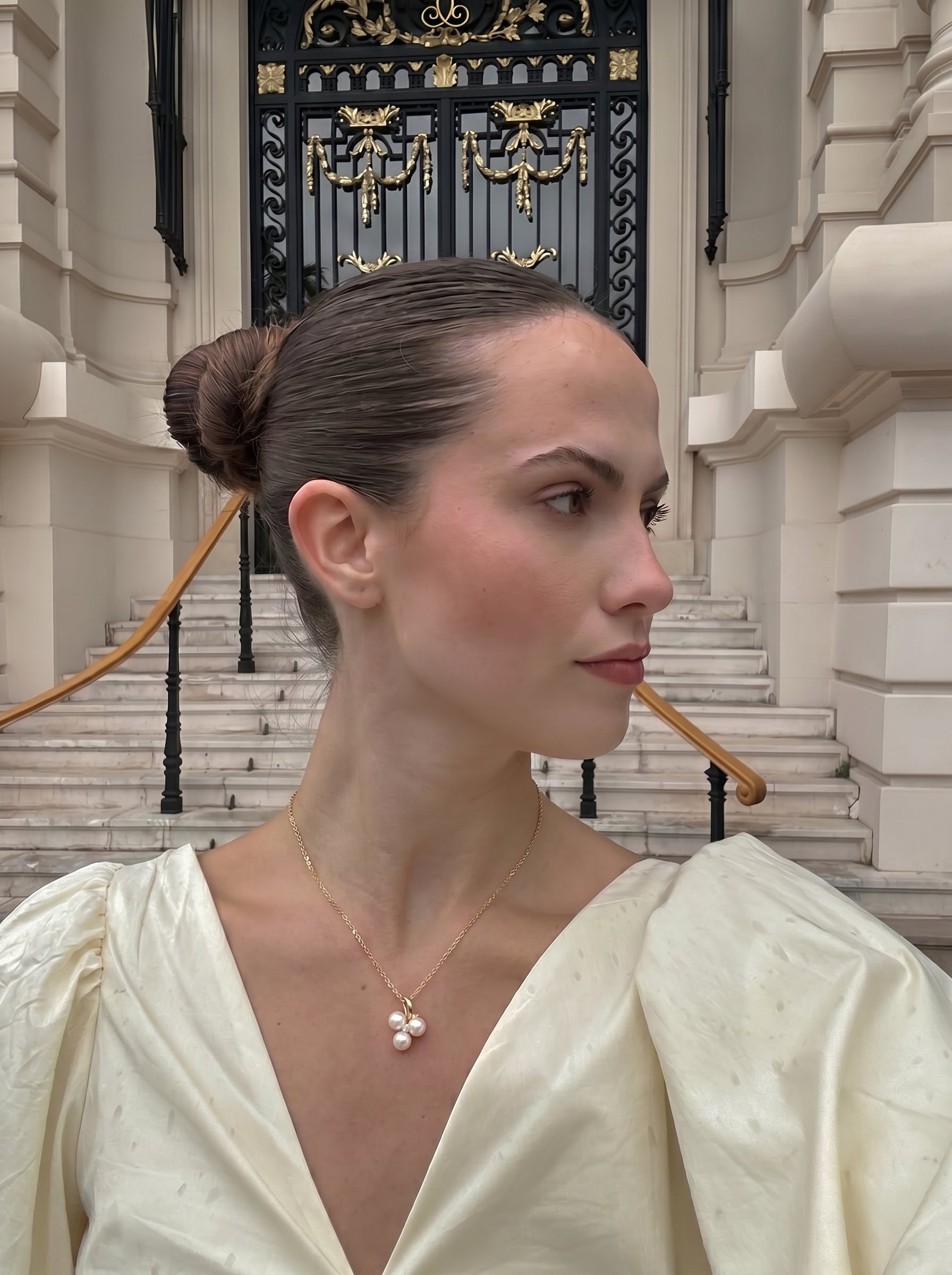 A woman in a cream dress with puffed sleeves wears the Trine Freshwater Pearl Trio Pendant Chain Necklace, featuring three small white pearls clustered on a gold chain, in front of a grand building with ornate doors and stairs. @lifestyle #lifestyle