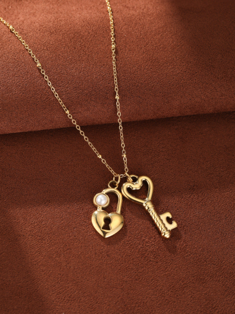 Collier ton or présentant des pendentifs cadenas et clé entrelacés en forme de cœur, le cadenas orné d'une perle blanche d'eau douce.