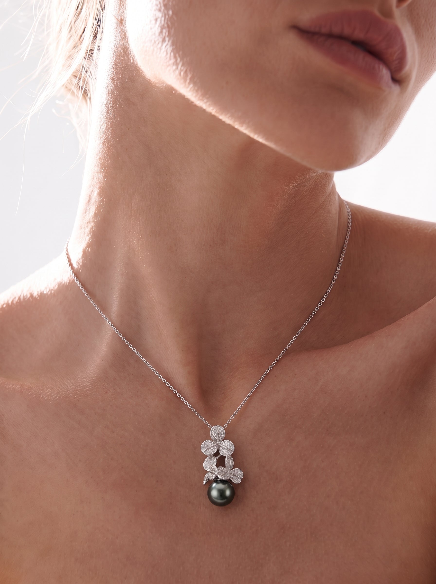 Close-up of a model wearing the Unfold Tahitian Pearl Clover Pendant Necklace, featuring a delicate silver chain and a pendant with clover-shaped motifs adorned with small stones, holding a single, lustrous black Tahitian pearl. @model