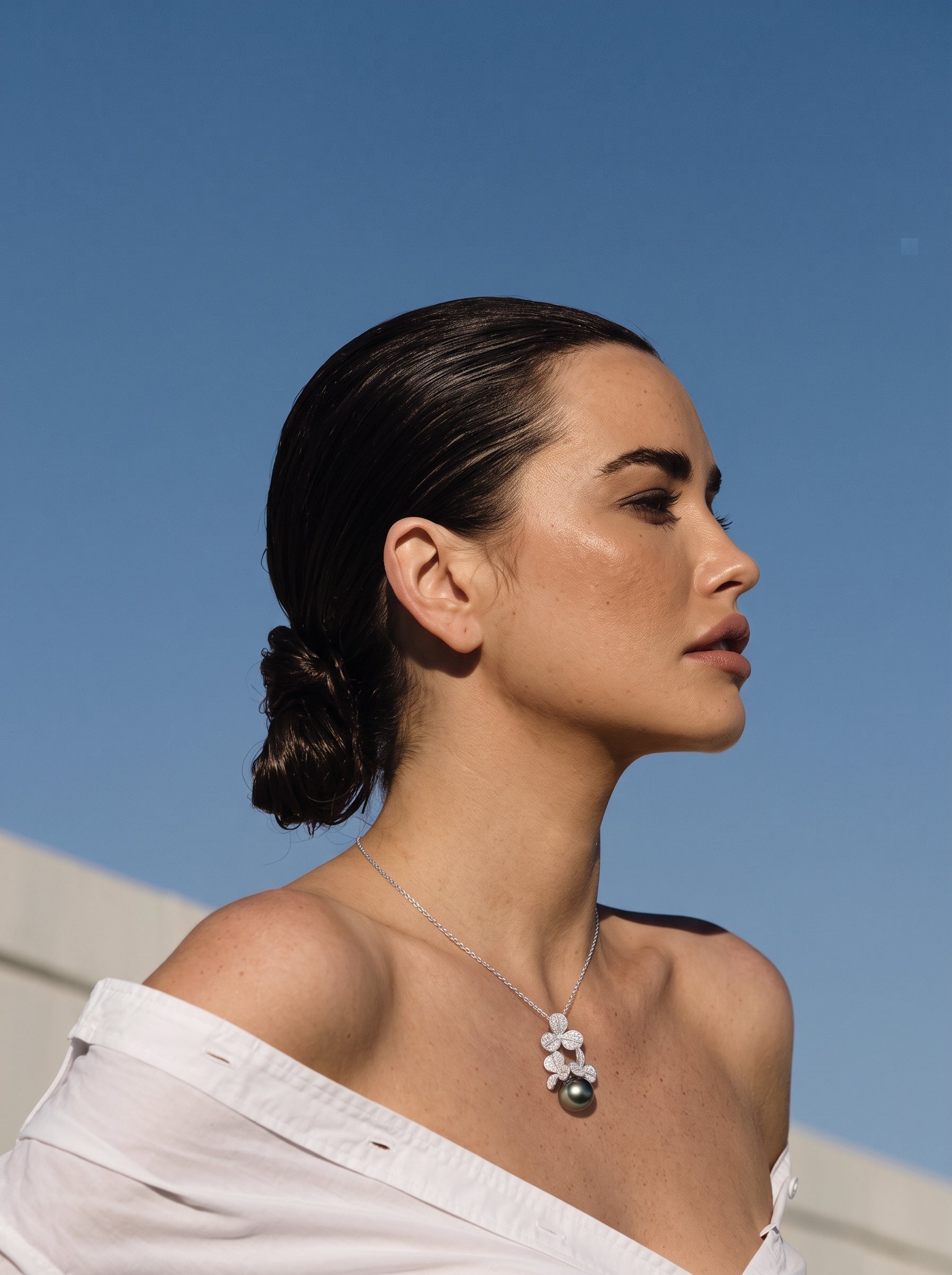 A woman wearing the Unfold Tahitian Pearl Clover Pendant Necklace, featuring a cluster of clover-shaped motifs adorned with sparkling stones and a large, dark Tahitian pearl, against a bright blue sky. @lifestyle
