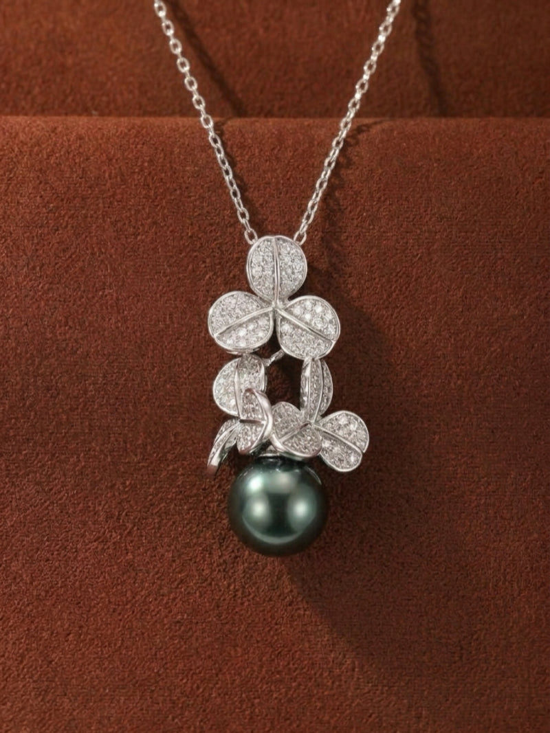 A delicate silver chain necklace with a pendant featuring a cluster of sparkling clover-shaped motifs cradling a lustrous, round 14mm Tahitian pearl in a deep black hue.