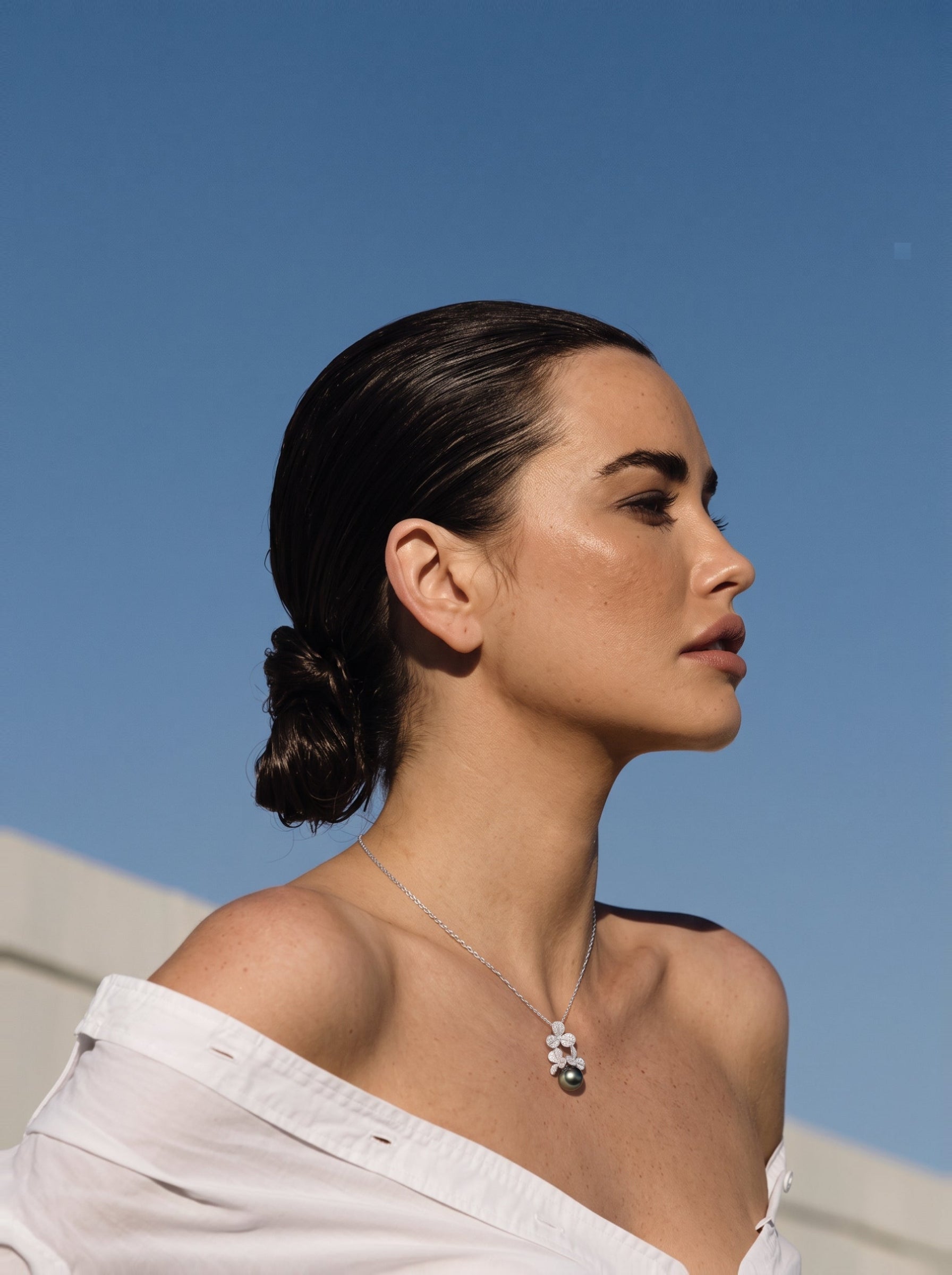 Une femme portant le collier pendentif trèfle Unfold en perle de Tahiti, présentant un groupe de motifs en forme de trèfle ornés de pierres scintillantes et une grande et sombre perle de Tahiti, contre un ciel bleu vif. @lifestyle