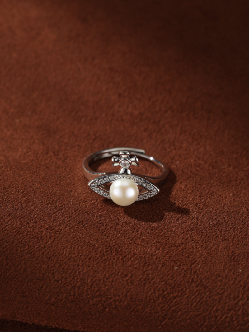 A silver cuff ring with a freshwater pearl, cross motif, and pave crystal details, set against a brown textured background.