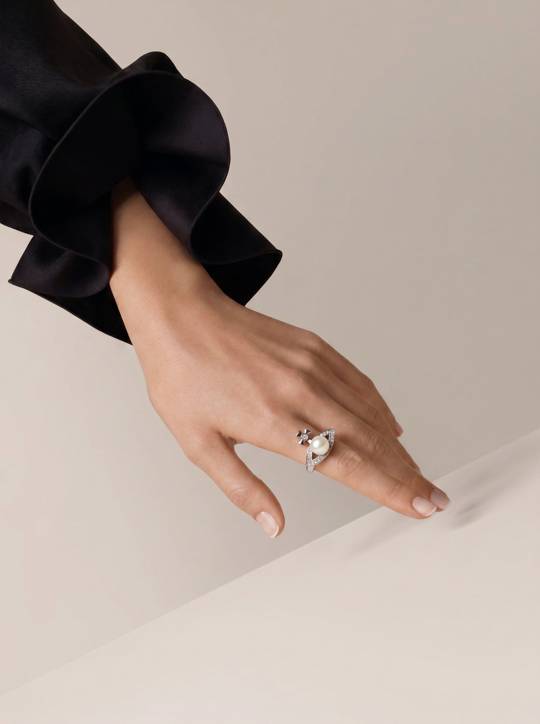 A woman's hand wearing a Vanguard Freshwater Pearl Cross Cuff Ring. The ring is silver-toned with a white pearl at its center, surrounded by a cross motif and pave details. The hand is resting on a surface, with a black ruffled sleeve visible.