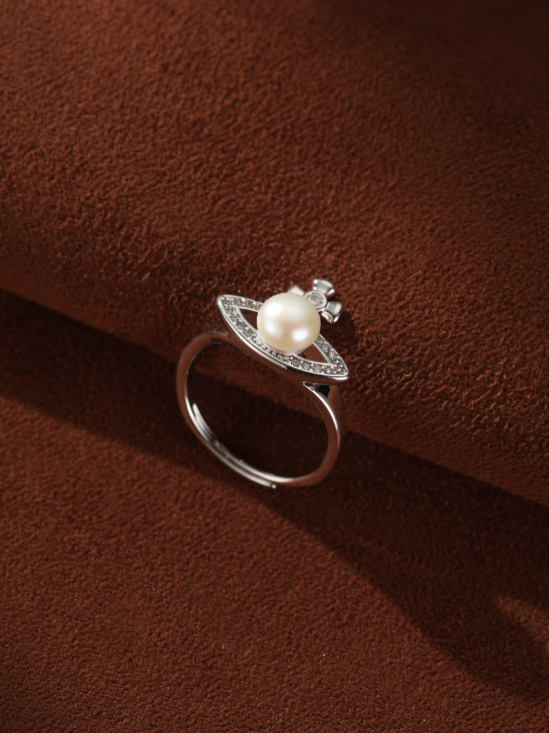 A silver-toned open cuff ring featuring a luminous freshwater pearl at its center, surrounded by a halo of pave-set stones that form an eye-like shape, with a subtle cross detail above the pearl.@pearl