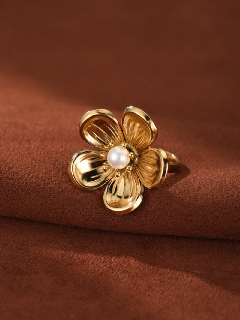 Gold-toned cuff ring shaped like a flower with a white freshwater pearl at its center, set against a brown textured background.