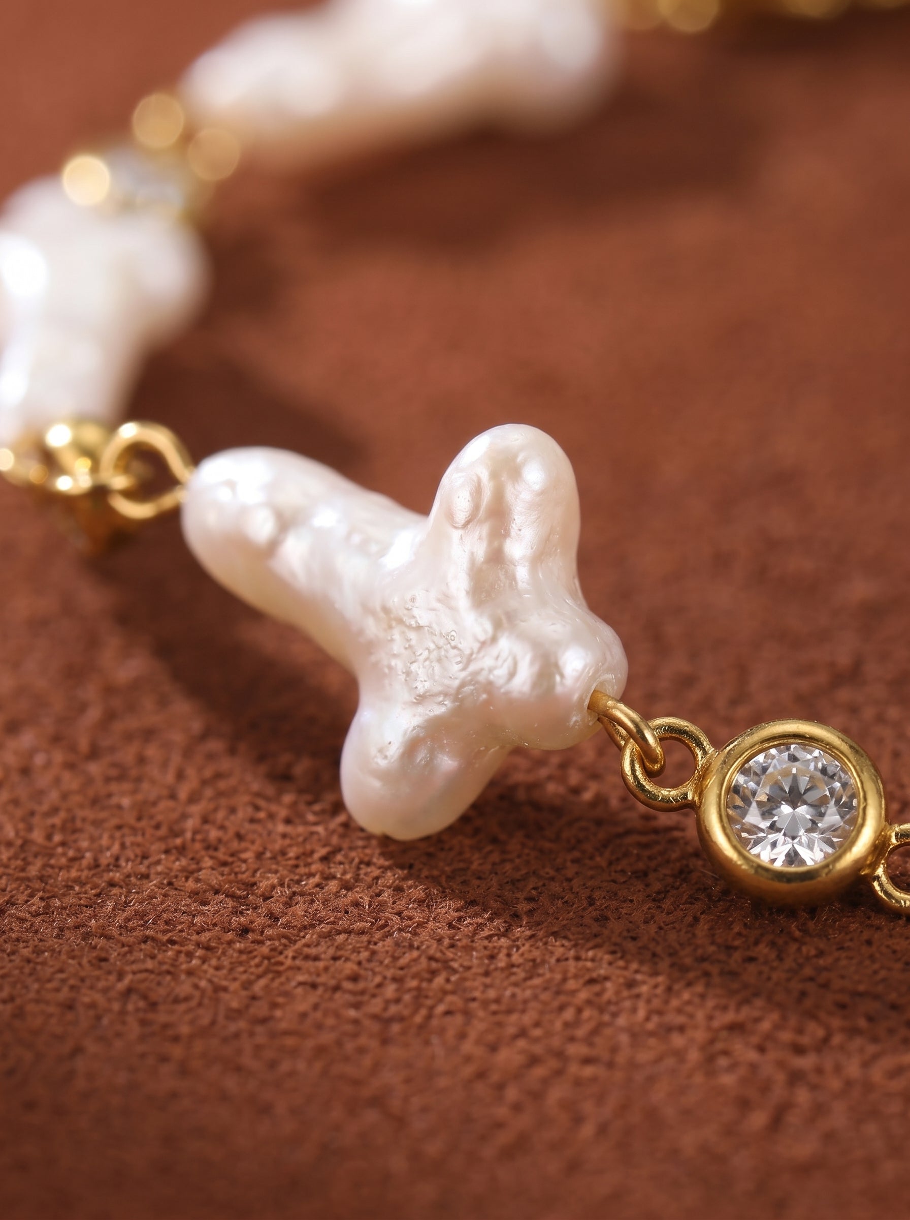 Close-up of a Verge freshwater pearl cross chain bracelet featuring a unique cross-shaped white baroque pearl with a gold chain and a clear crystal accent on a brown textured background.