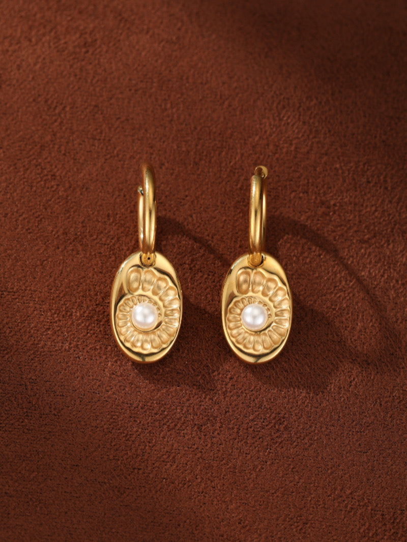 Gold spiral embossed oval drop earrings featuring a white freshwater button pearl at the center, set against a textured brown background.