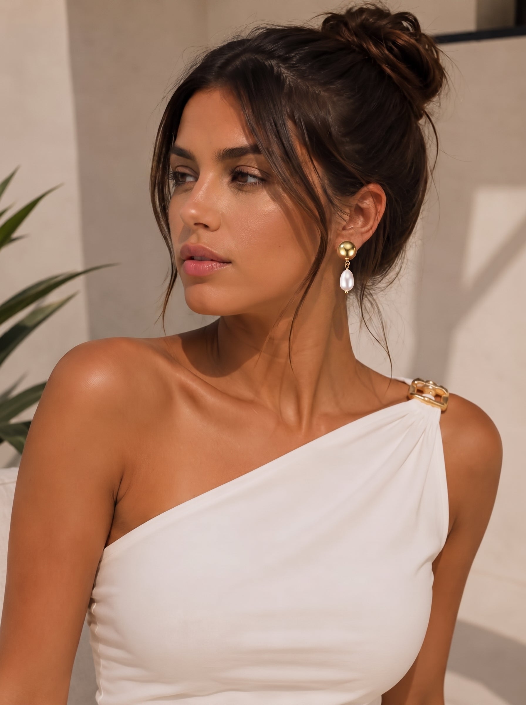 Une femme aux cheveux relevés en chignon porte les boucles d'oreilles pendantes Vesper en perle d'eau douce, qui présentent une sphère dorée et une perle blanche pendante, associées à un haut blanc à une épaule. @lifestyle