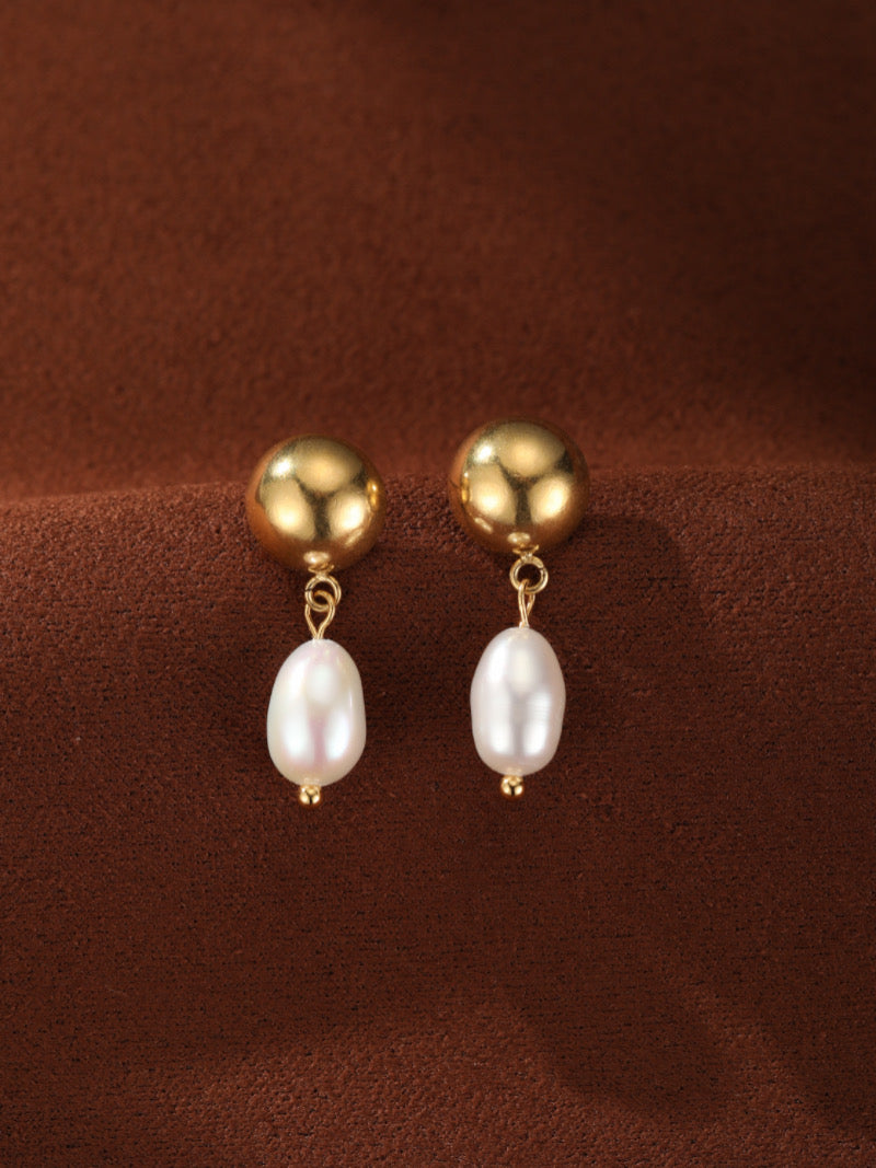 Boucles d'oreilles goutte en perle d'eau douce Vesper avec sphère ton or et perle blanche d'eau douce sur fond texturé marron.